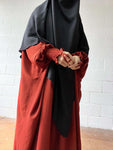 A close-up of a woman wearing a red Butterfly Abaya with smocked sleeves, her hands clasped in front, and a black headscarf.
